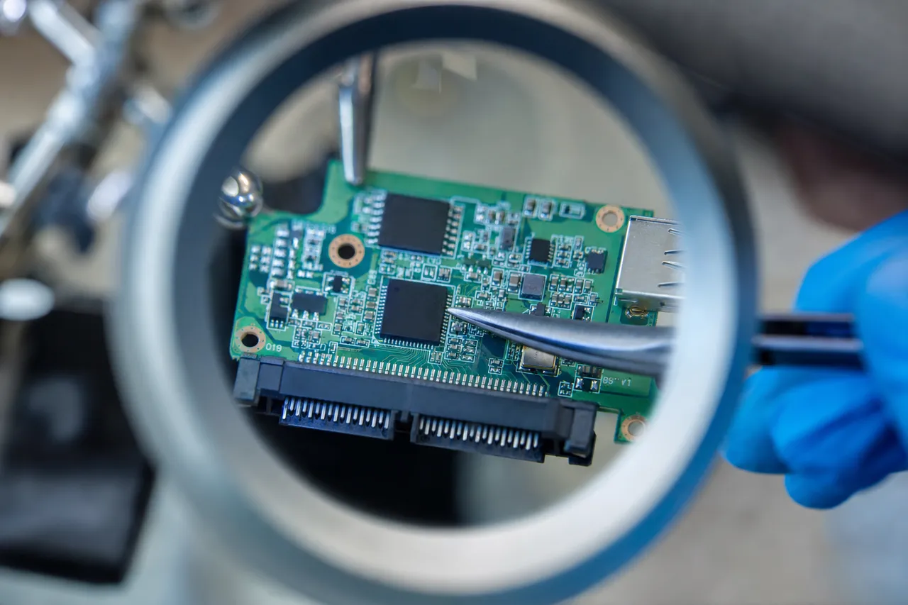 A green USB bridge circuit board from a portable external drive viewed under a magnifier, showing the USB connector, controller chips, and the SATA pin header, with anti-static tweezers held in a blue-gloved hand
